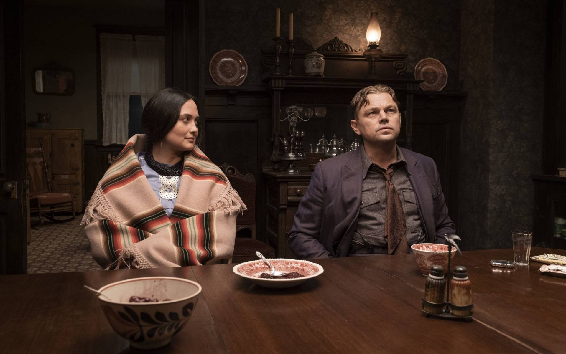 An Indigenous woman and a white man in a suit sit at a heavy wood dining table. China cabinets line the wall behind them.