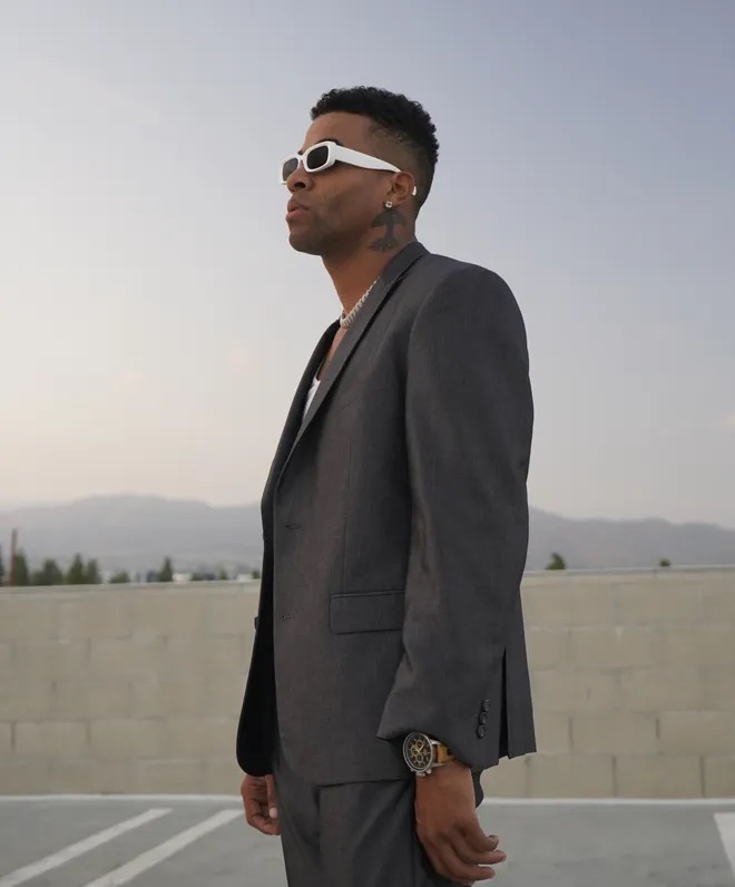 Konkrete looks into the distance while wearing a grey suit and sunglasses. His oak tree neck tattoo, symbolizing Oakland, is visible.