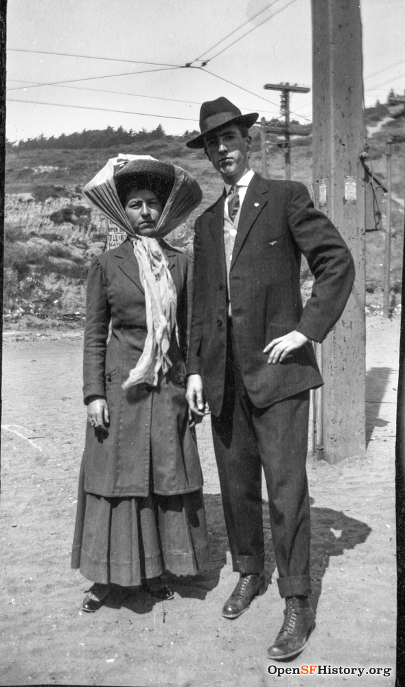 A Victorian couple stands on the side of a road. 