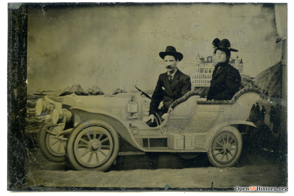 A man and a woman pose inside a prop car in a photo studio. Turn of the century.