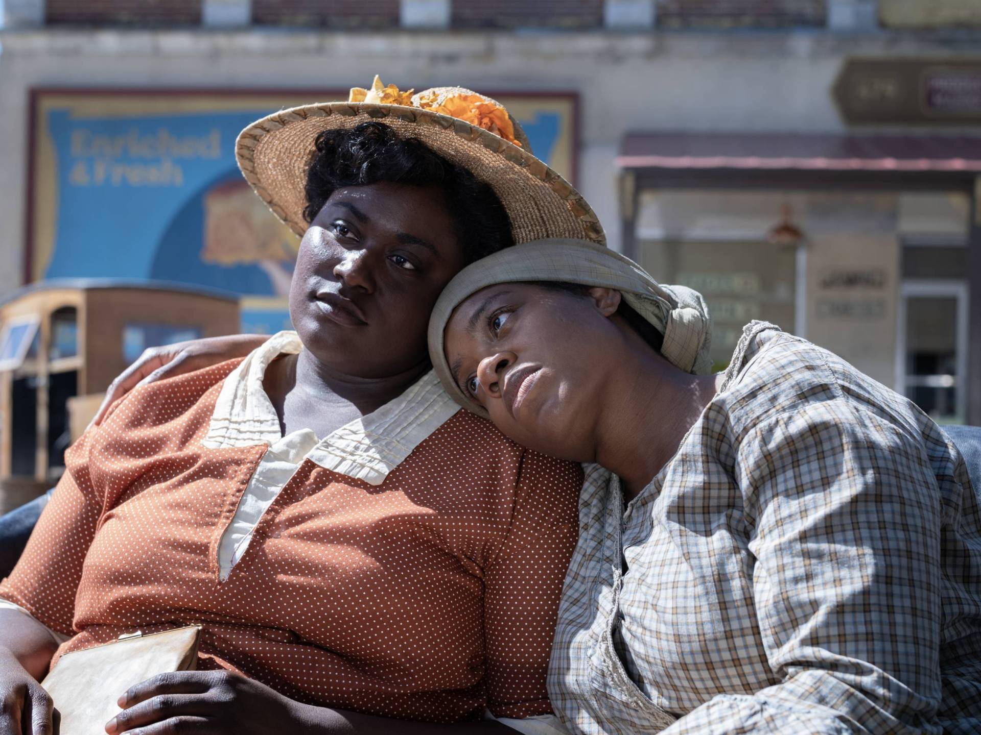 Two Black women sit outdoors in the sun, their heads leaning on one another. They look dejected.