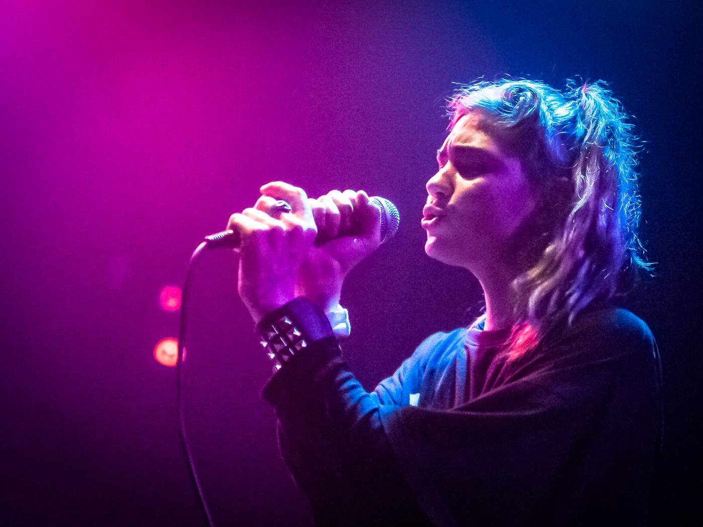 Person holds mic with both hands and sings with eyes closed under pink and purple light