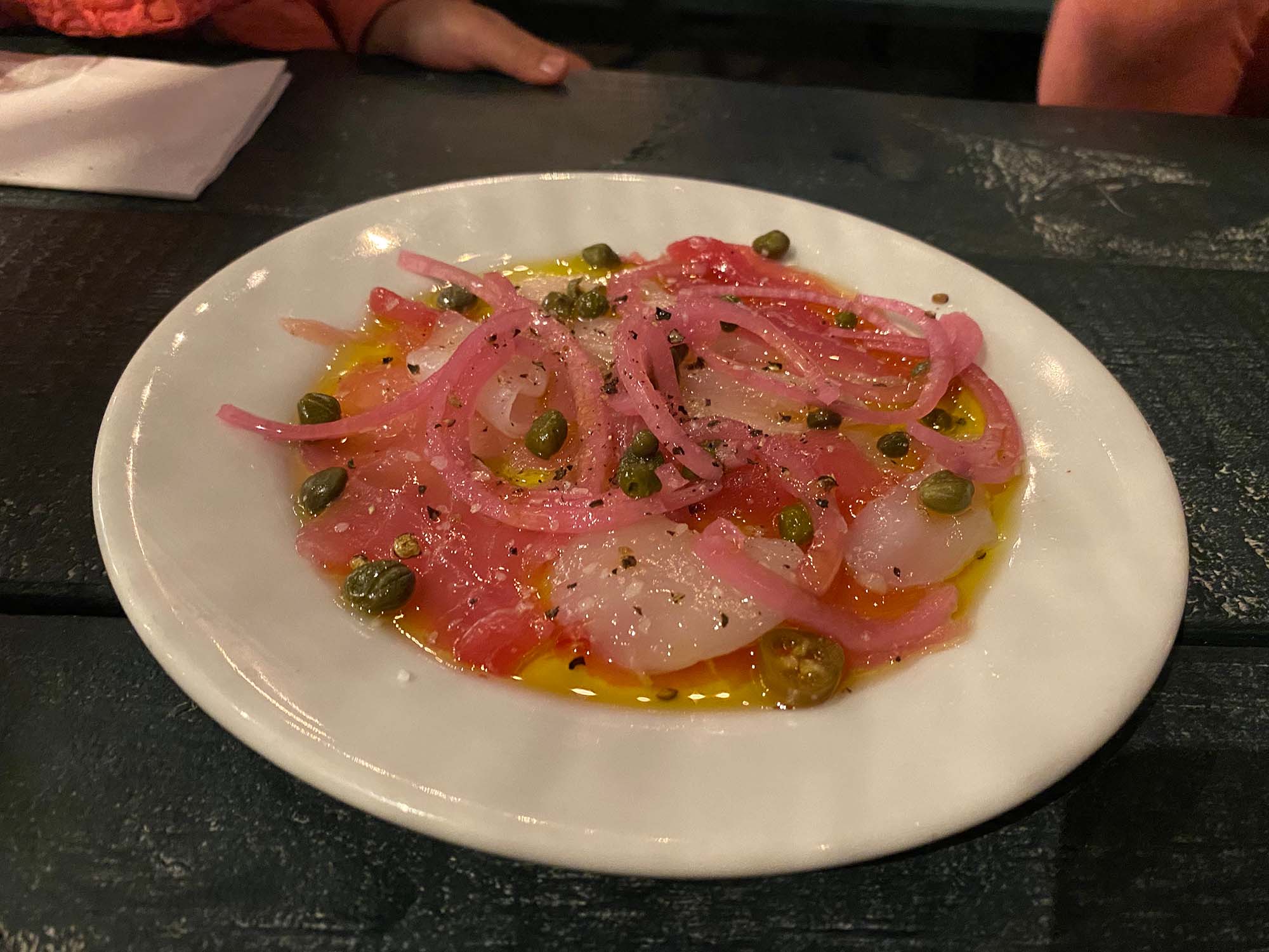 Seafood crudo topped with capers and red onions, served in a shallow white bowl.