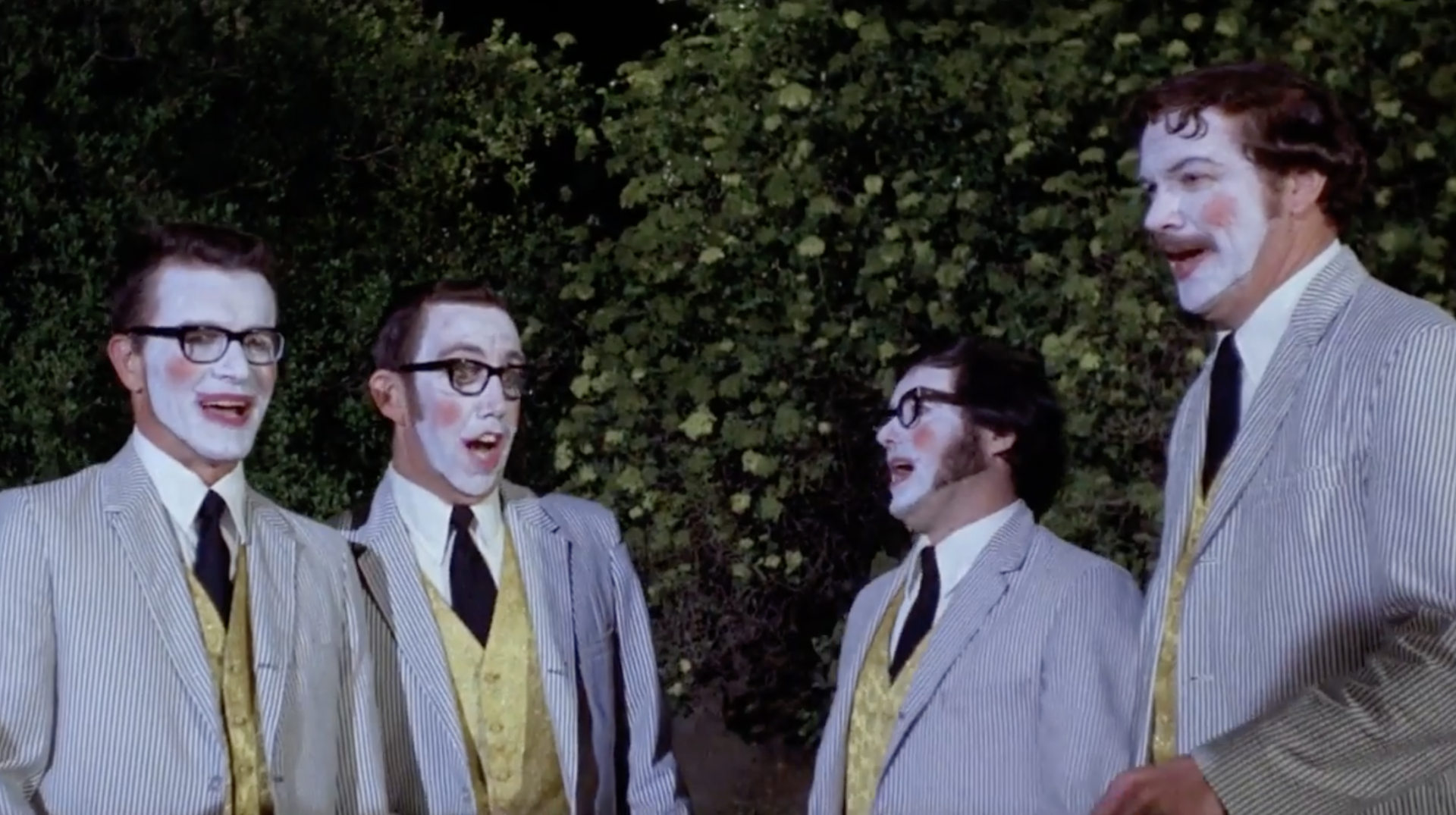 Four men in matching facepaint and grey and yellow suits sing together. They are all wearing glasses.