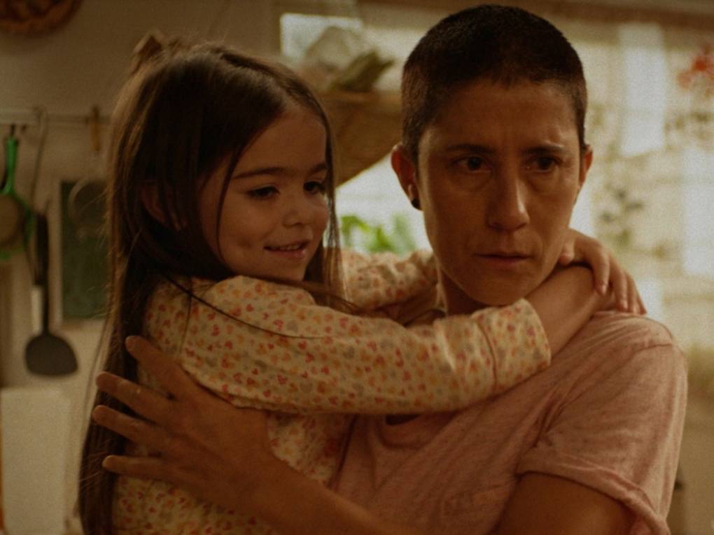 A sickly looking person with a shaved head holds a small girl. The child is smiling.