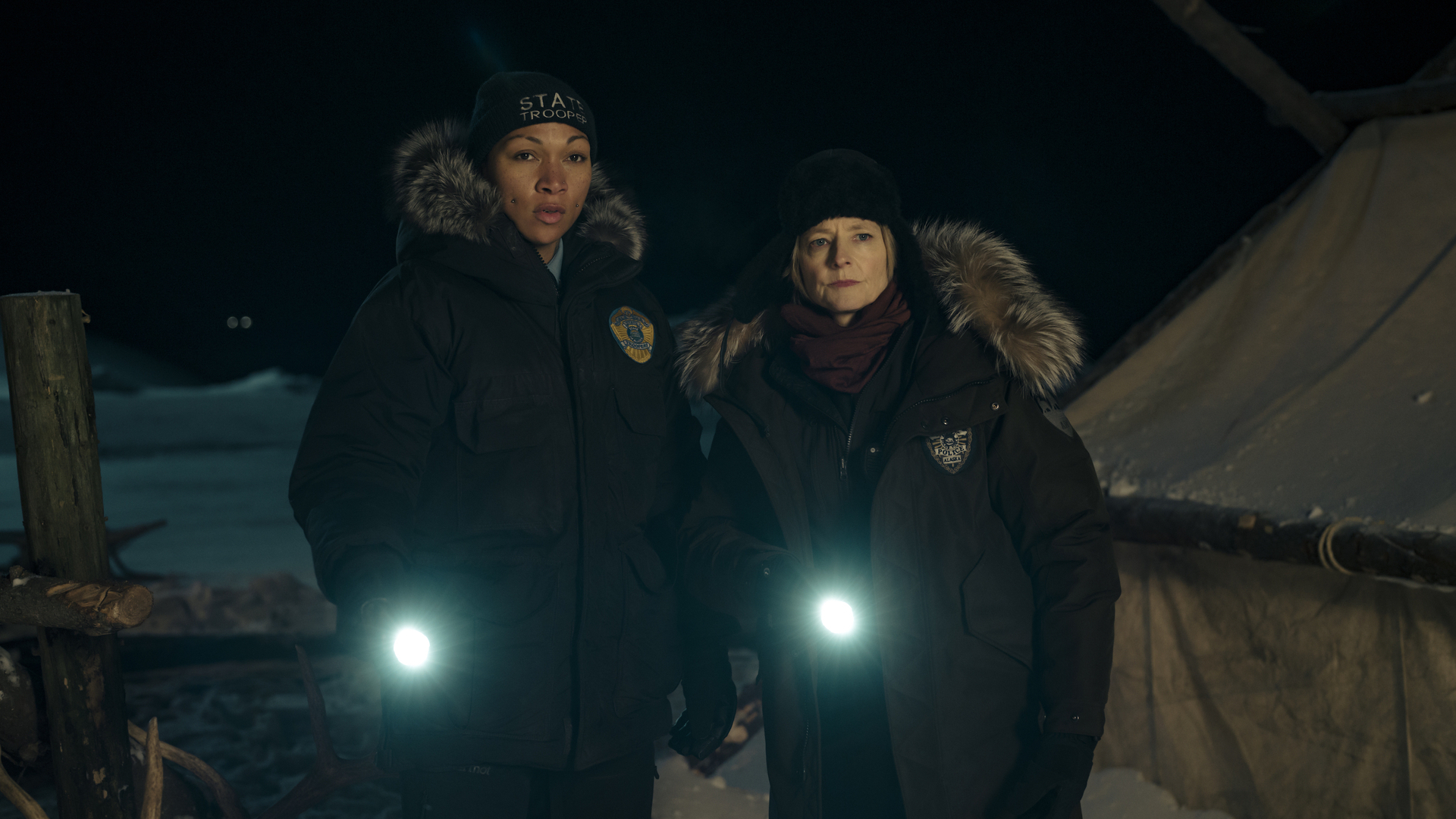 Two female police officers stand side-by-side wearing large padded winter coats and shining flashlights into the distance.
