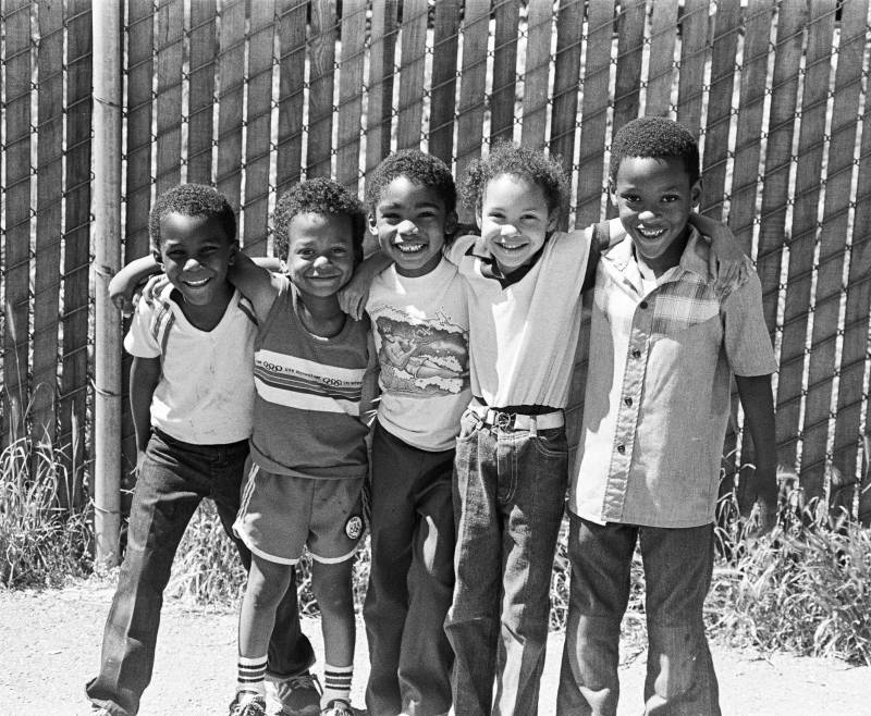 An archival photo of bright smiles radiating from young Black students against a wood-and-chainlink fence.