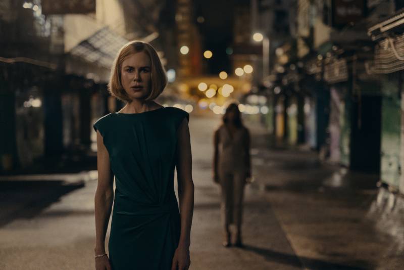 A slender middle aged white woman stands in a quiet dimly lit street. A figure lurks behind her.