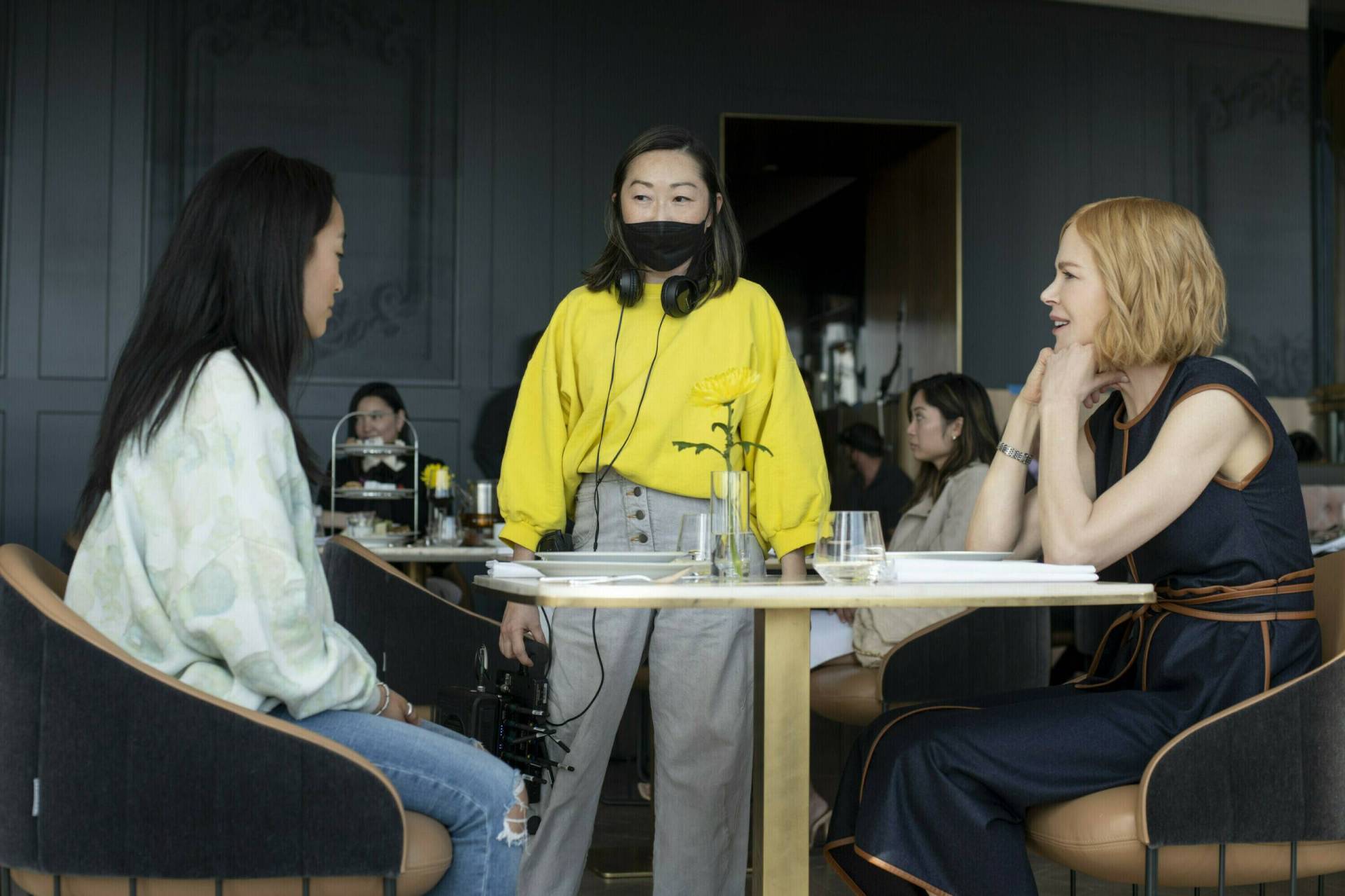 A white woman and an Asian woman sit across from each other in a restaurant. A woman wearing a mask and headphones around her neck stands between them.