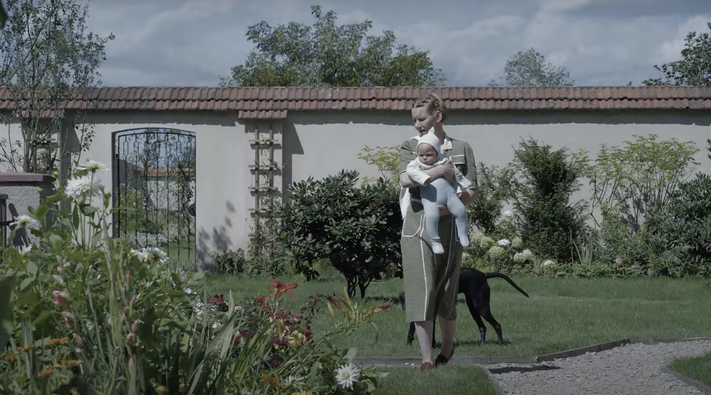 A woman in 1940-era clothing stands in a walled garden clutching a baby. A dog stands behind them.