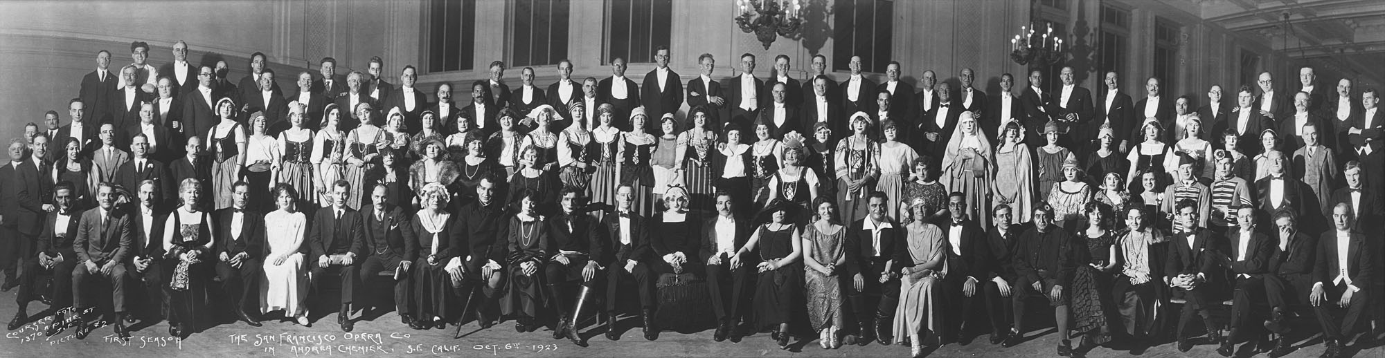 Large black-and-white group photo of people in costumes and tuxes on risers inside high-ceilinged space