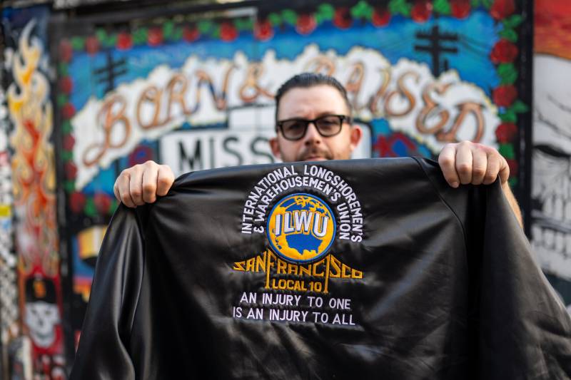 A person wearing sunglasses holds up a jacket with "International Longshoremen's Warehousemen's Union" written on it.