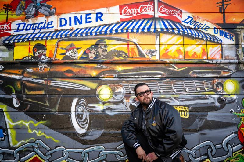 George Crampton Glassanos posing in front of his mural of a black droptop lowrider and the Mission staple, Doggie Diner.