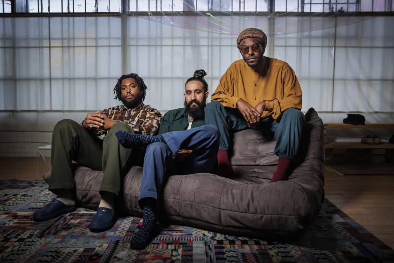 Three men sit on a brown couch looking straight at the camera.