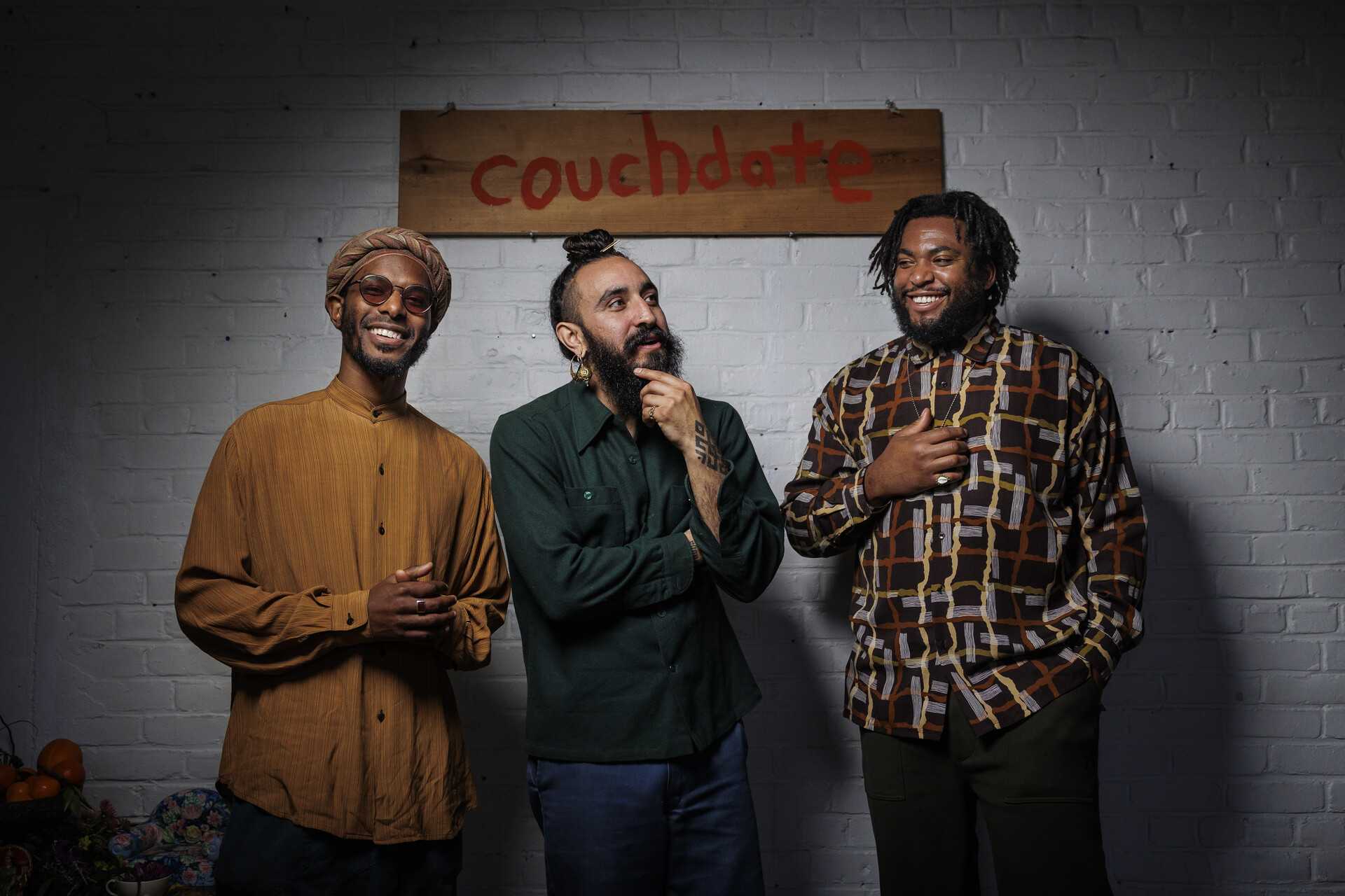 Three men stand in front a white wall that has a sign that reads "couchdate" on it.
