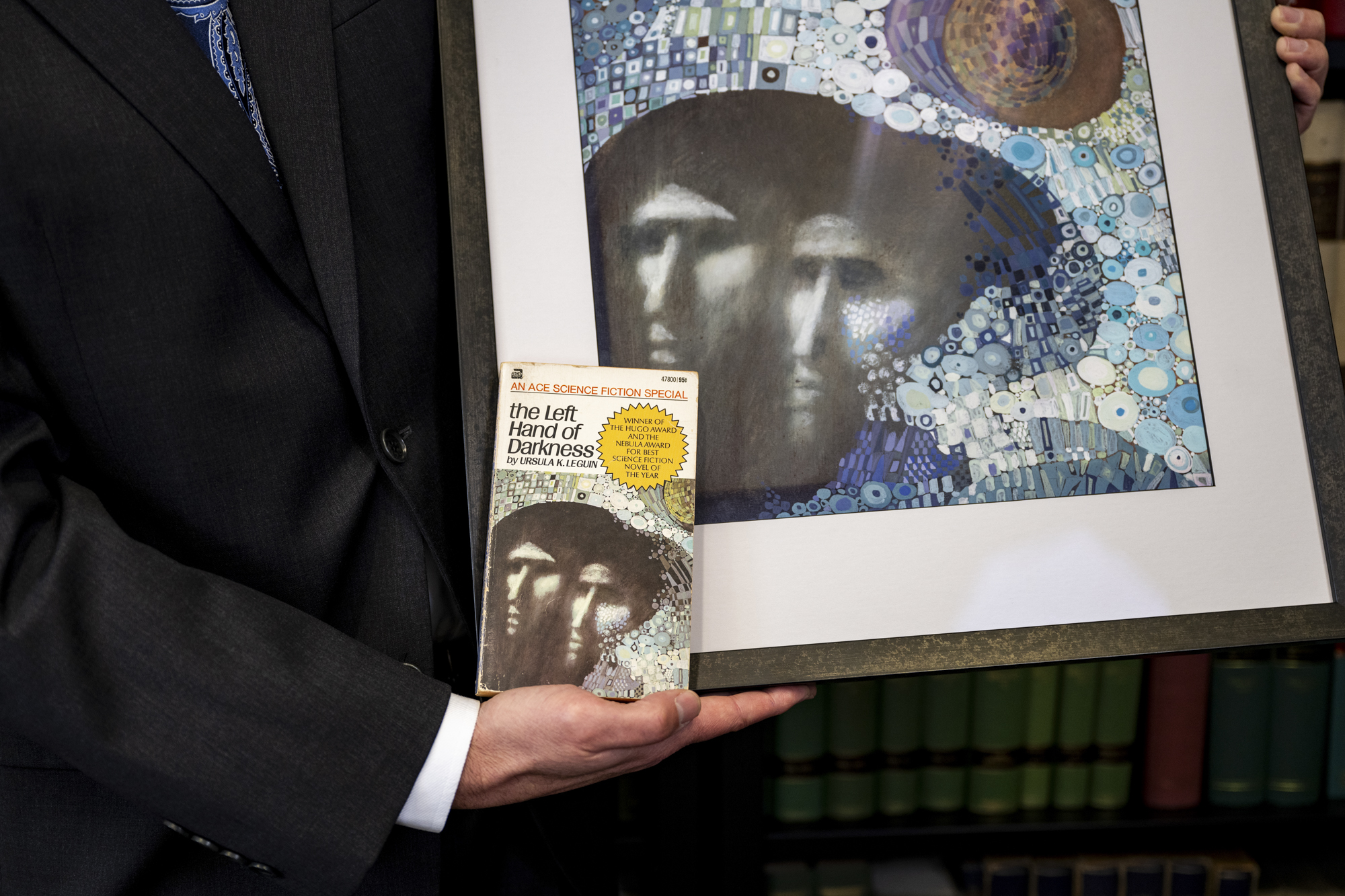 Hand holds a paperback book in front of a framed painting with the larger image of the book cover: two blurry faces against an abstract field