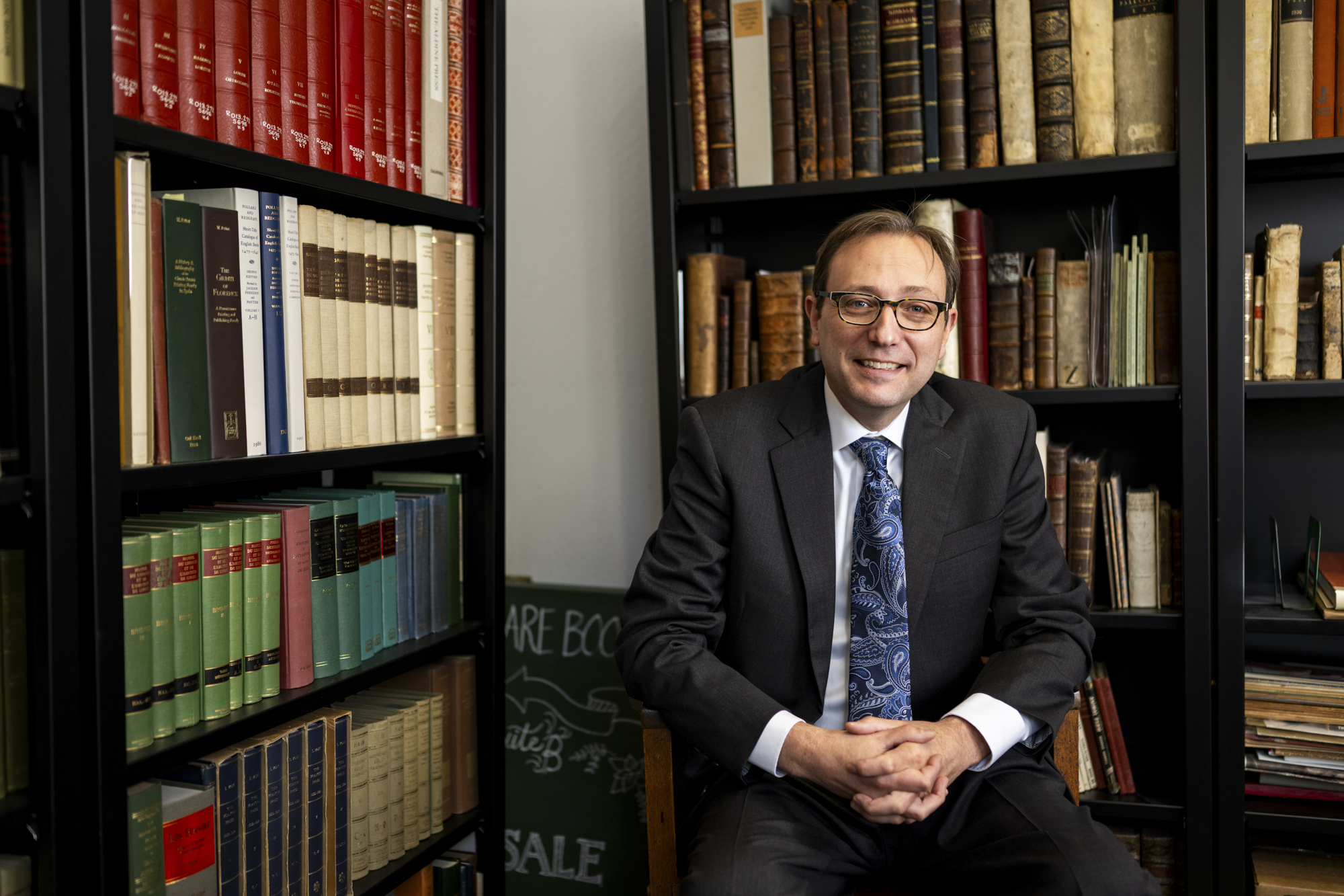 Person in suit with glasses smiles and sits in front of full bookshelves