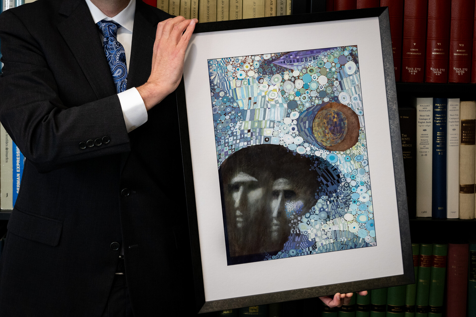 Person in suit holds a framed painting of two people in an abstracted landscape