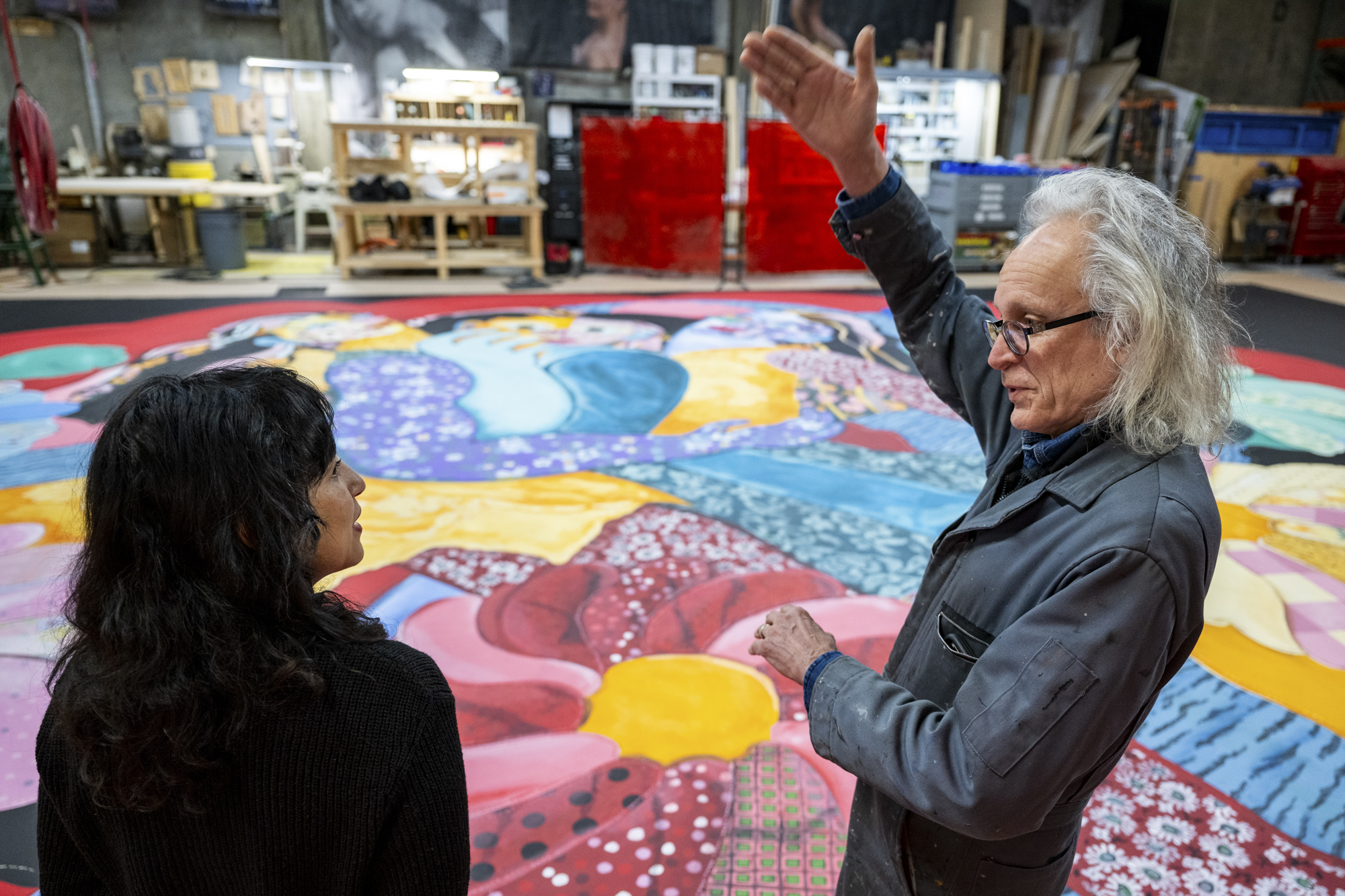 Two people talk, one gestures with arms in front of brightly painted large canvas on floor