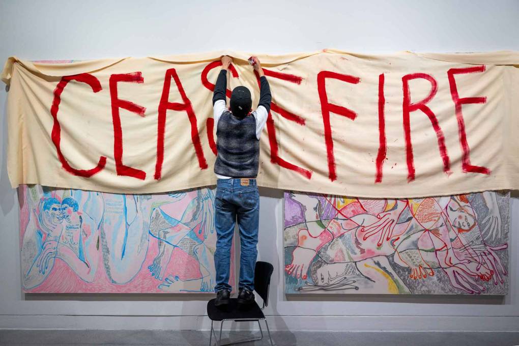 a man in jeans hangs a sign reading Ceasefire over two paintings against a white wall