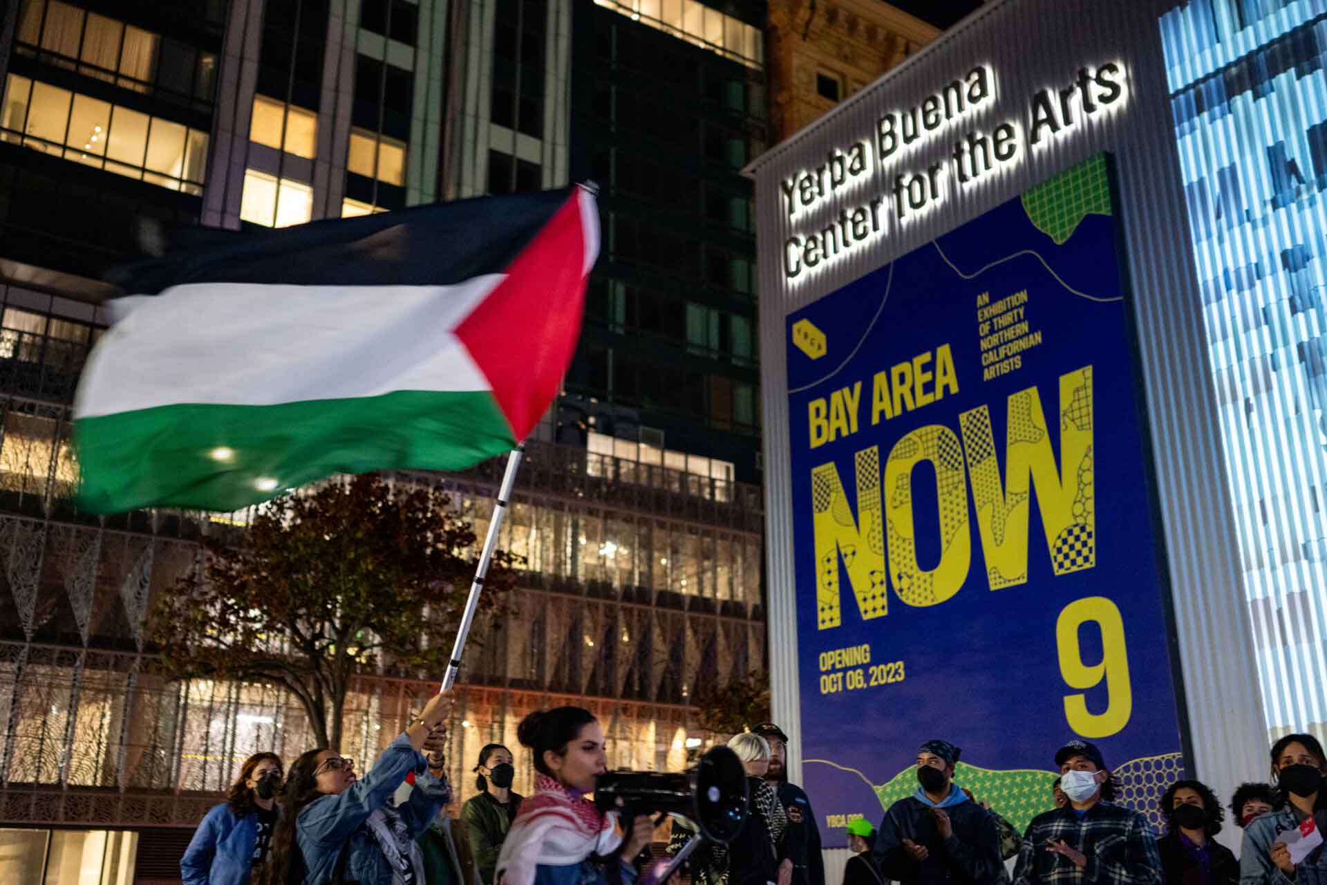 A woman in a denim jacket waves a Palestinian flag outside a museum building at night