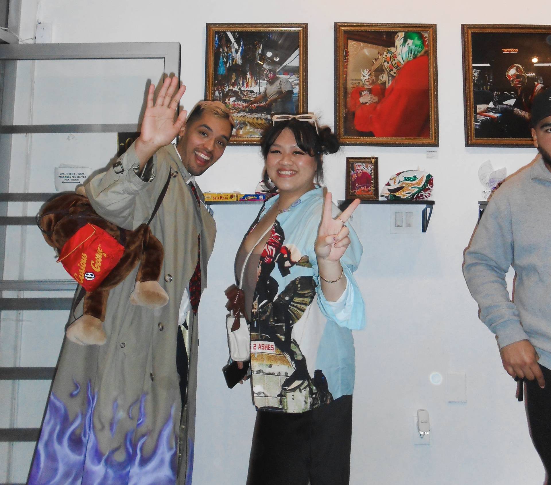 Two people wearing colorful clothing wave at the camera while posing in front of an art exhibit.