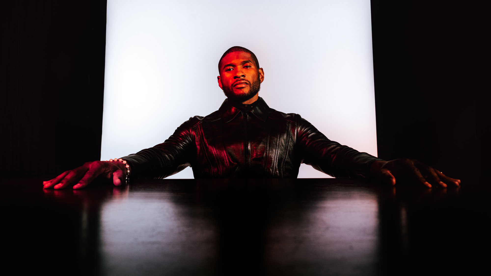 Person lit red in black jacket sits with arms out over dark table