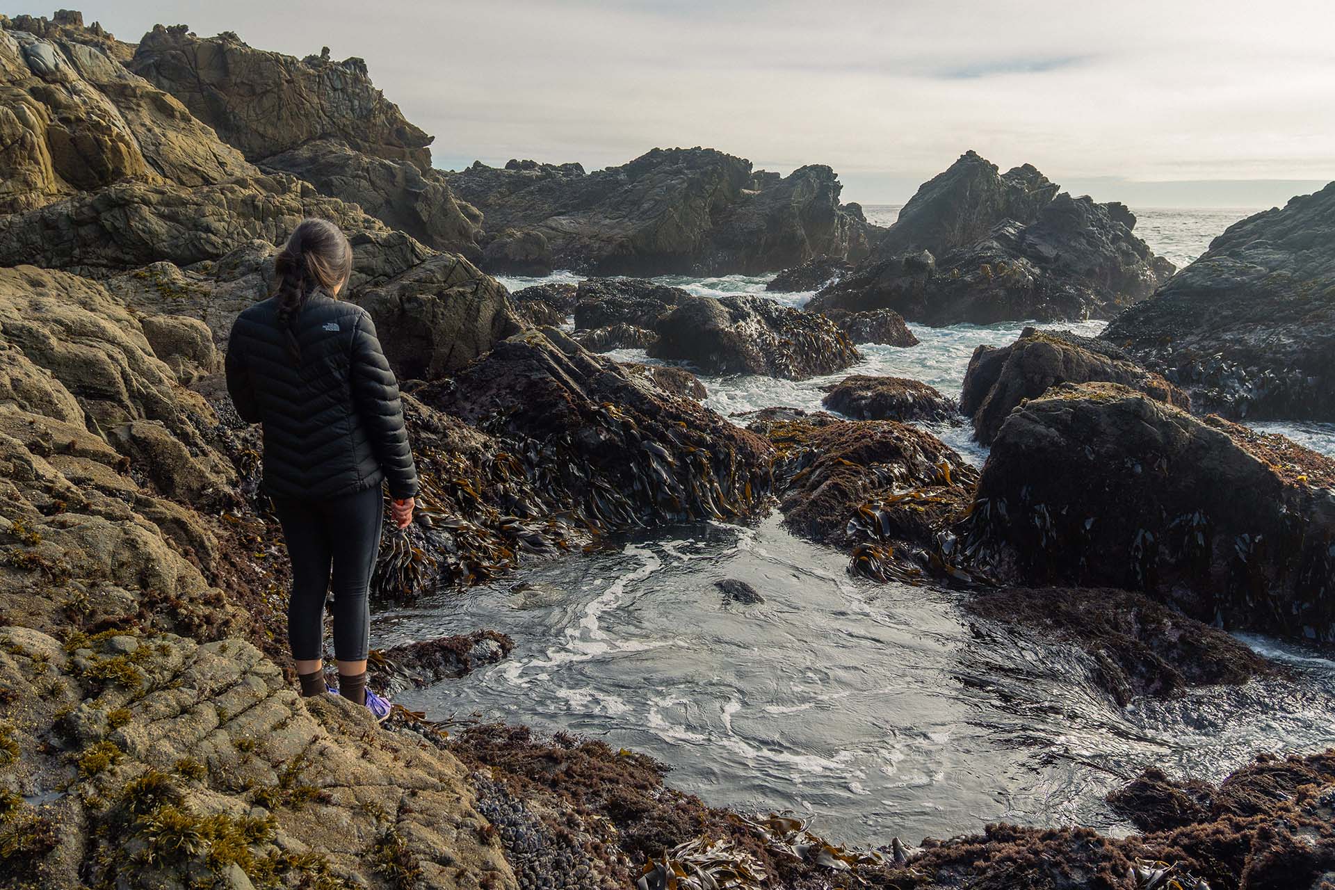 A woman in black stands at the edge of a tidepool looking down toward the water.