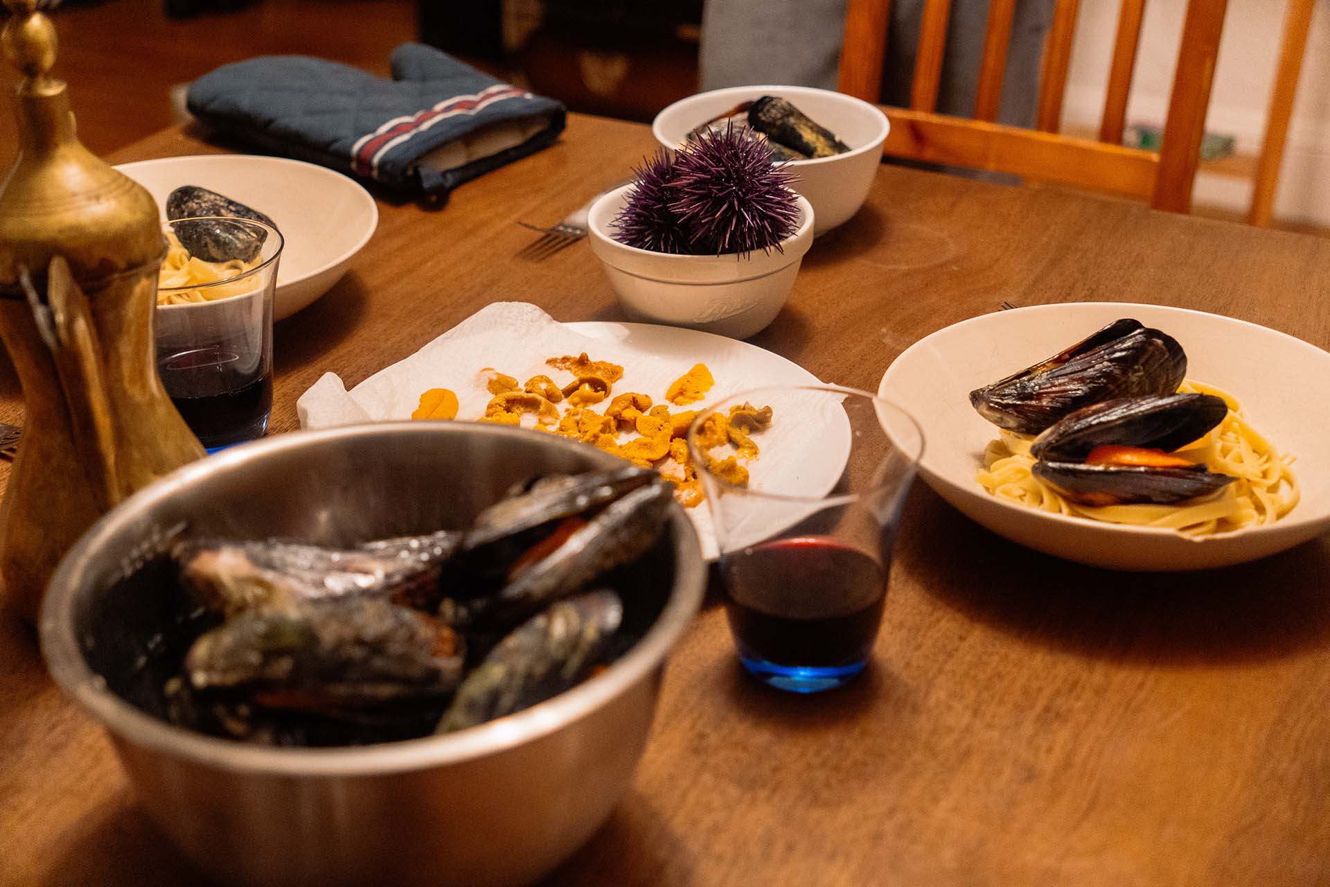 Plates of pasta, mussels and fresh uni laid out on a dining table with glasses of red wine.