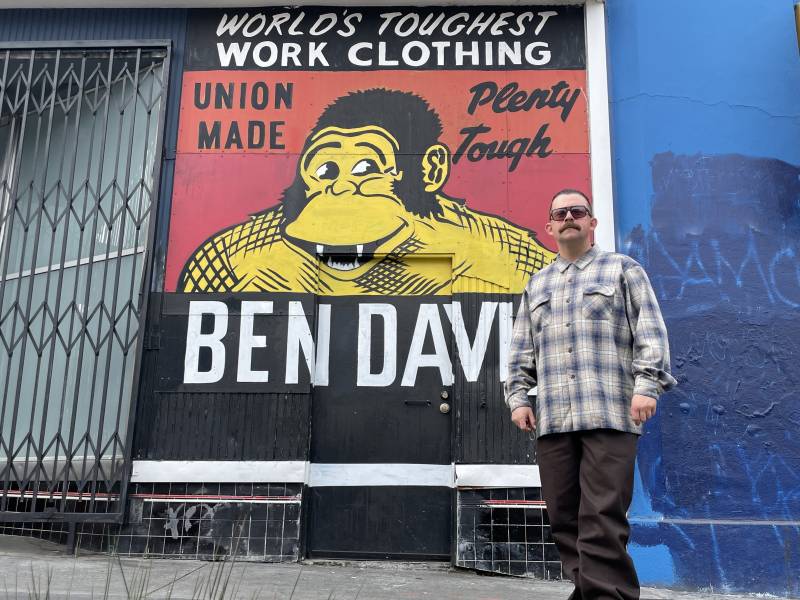 Person in plaid button-up shirt standing in front of the Ben Davis sign, where the iconic logo of a yellow gorilla with a charismatic smile serves as the main image, and the words "World's Toughest Work Clothing" sits atop the sign. 