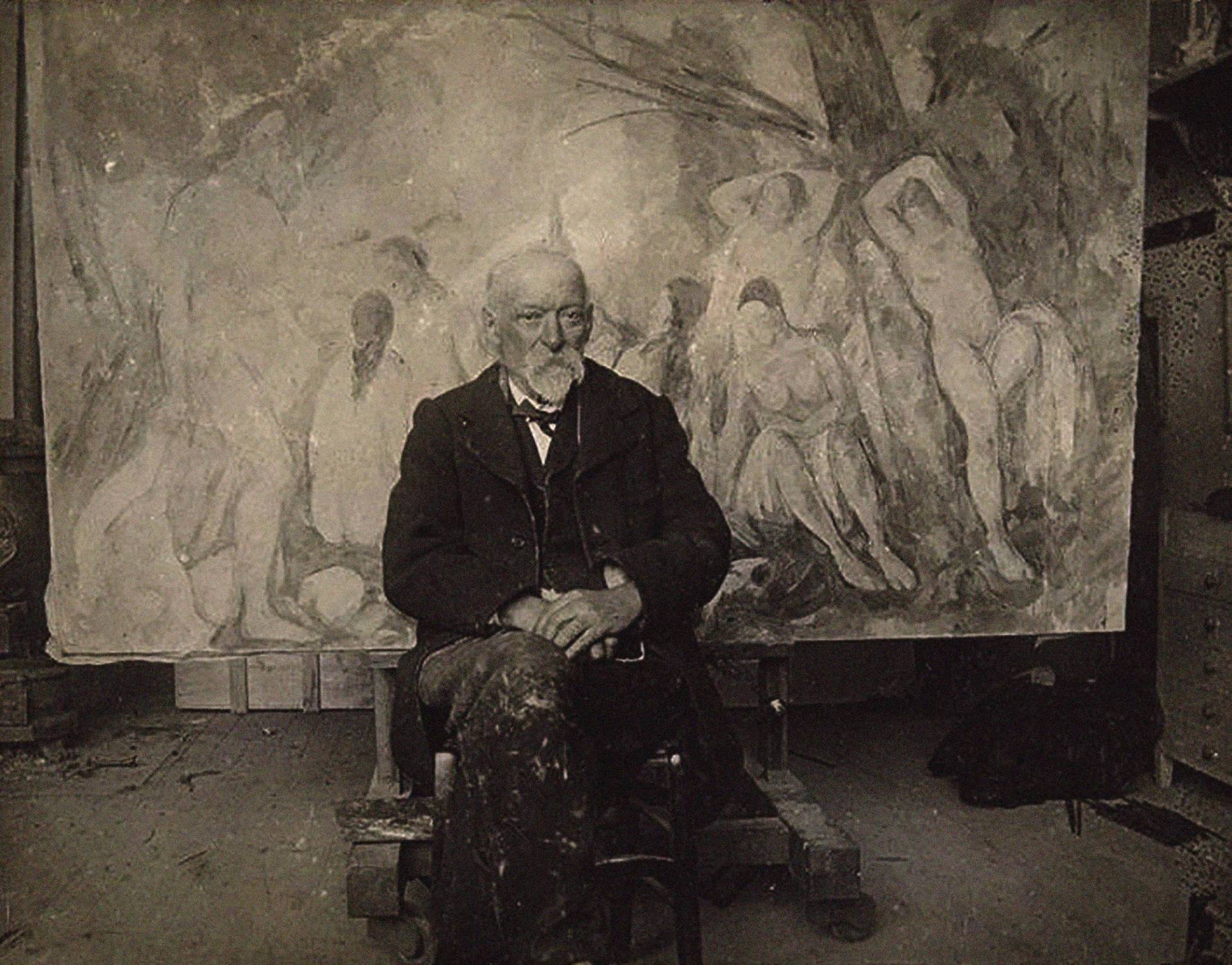 An elderly man with white beard in a suit sits in front of a painting of people and trees.