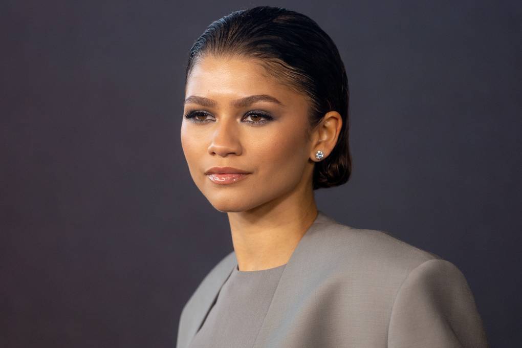 A young Black woman stands before a dark grey wall wearing a light grey suit and diamond earrings.