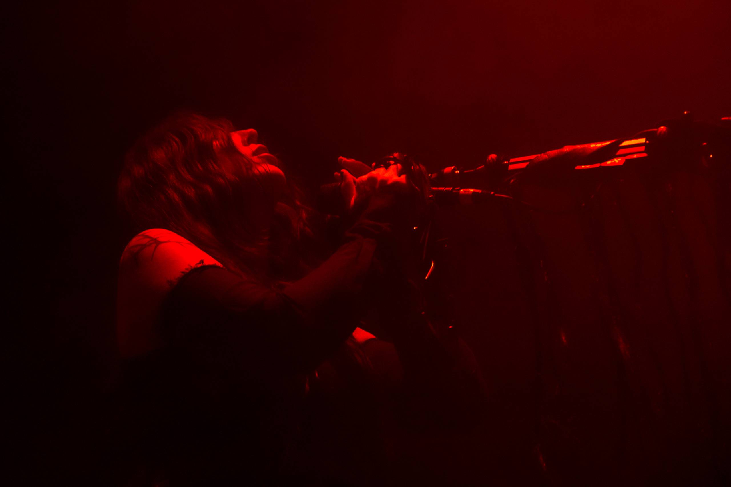A woman tilts her head back while holding onto a microphone. She is dimly lit in red light.