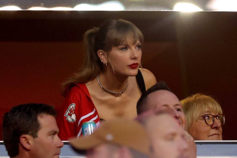Taylor Swift is photographed leaning forward in a crowd, wearing a red football jersey and looking puzzled.