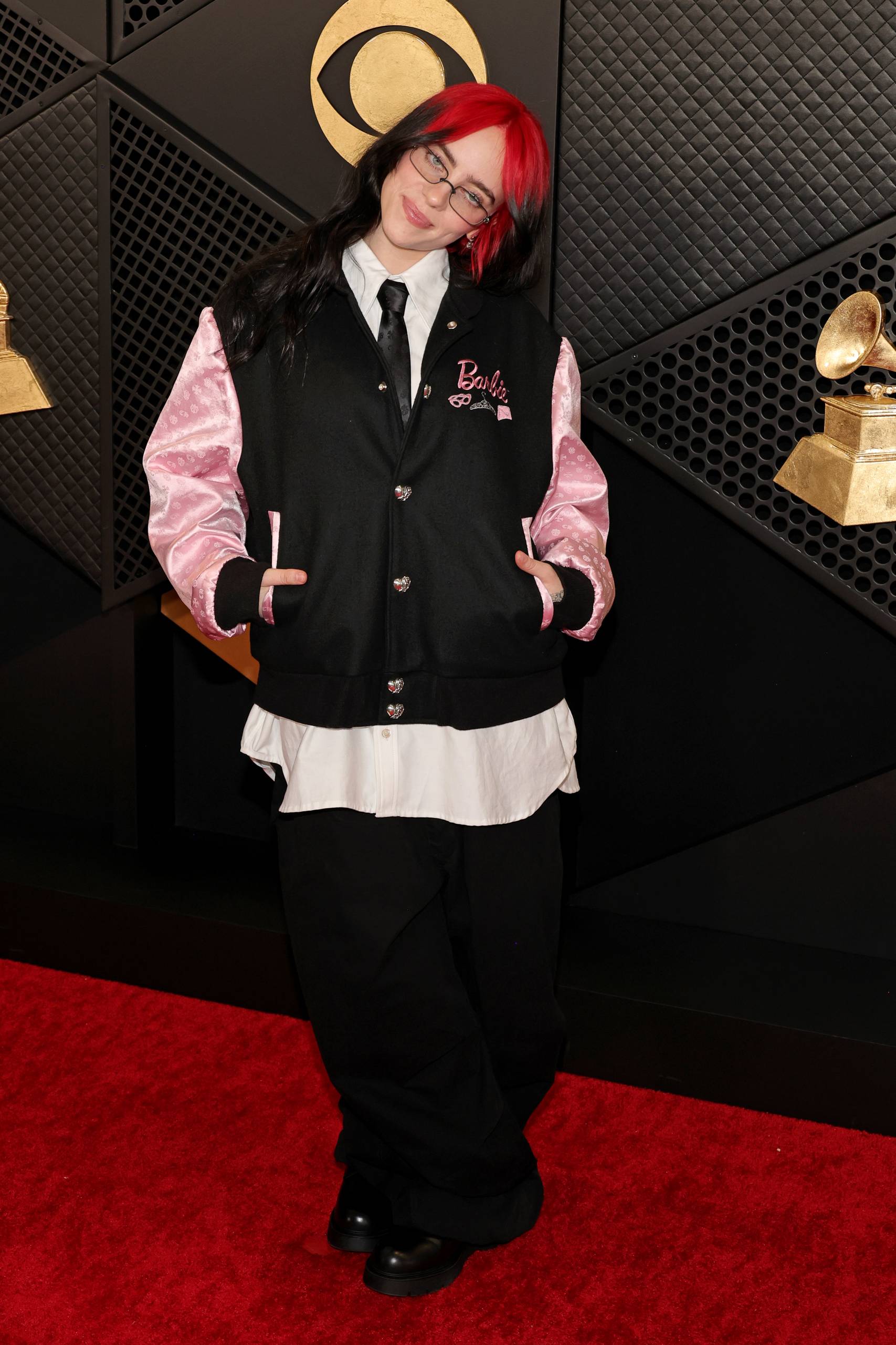A young woman with red and black hair stands on the red carpet wearing oversized black pants and a bomber jacket.