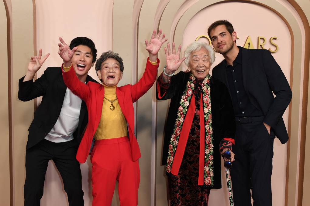 A young Asian man and two senior Asian women gesture wildly on a red carpet. A second man stands to their right.