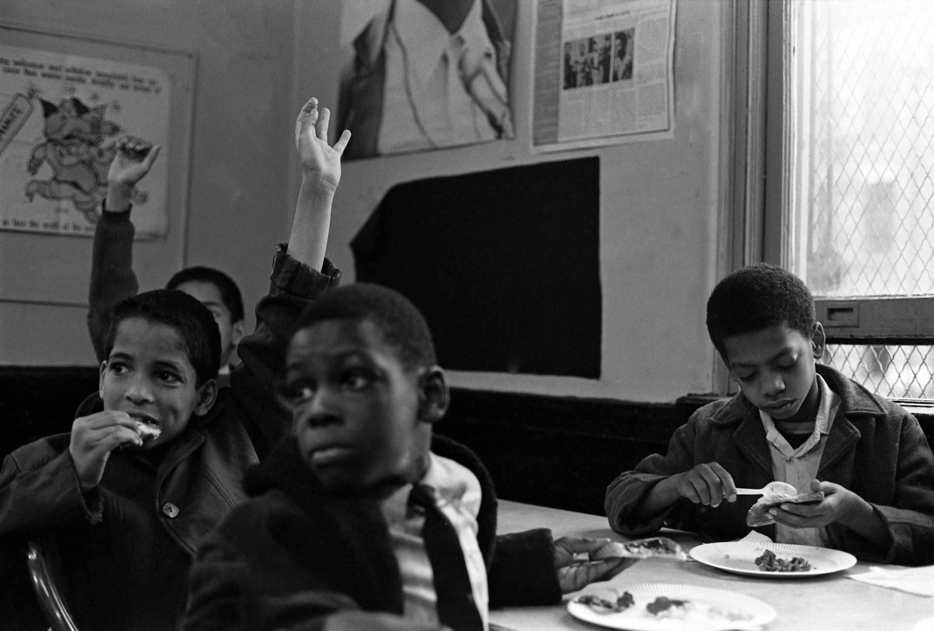 Several young Black boys, one of whom is wearing a suit, raise their hands to speak as they sit around a table, paper plates of food in front of them.