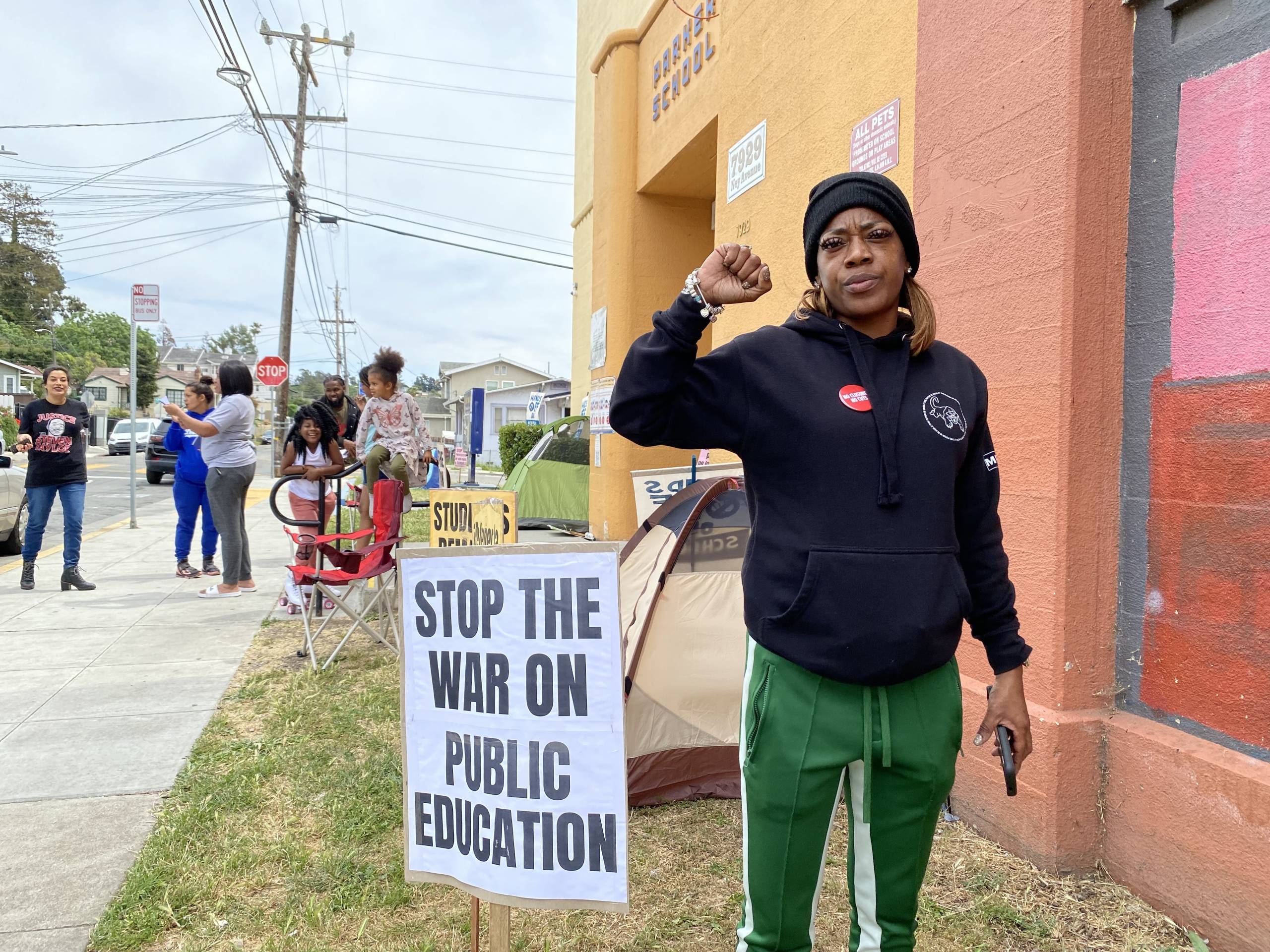 A woman stands outside a building with her fist up. A sign next to her says "stop the war on public education."