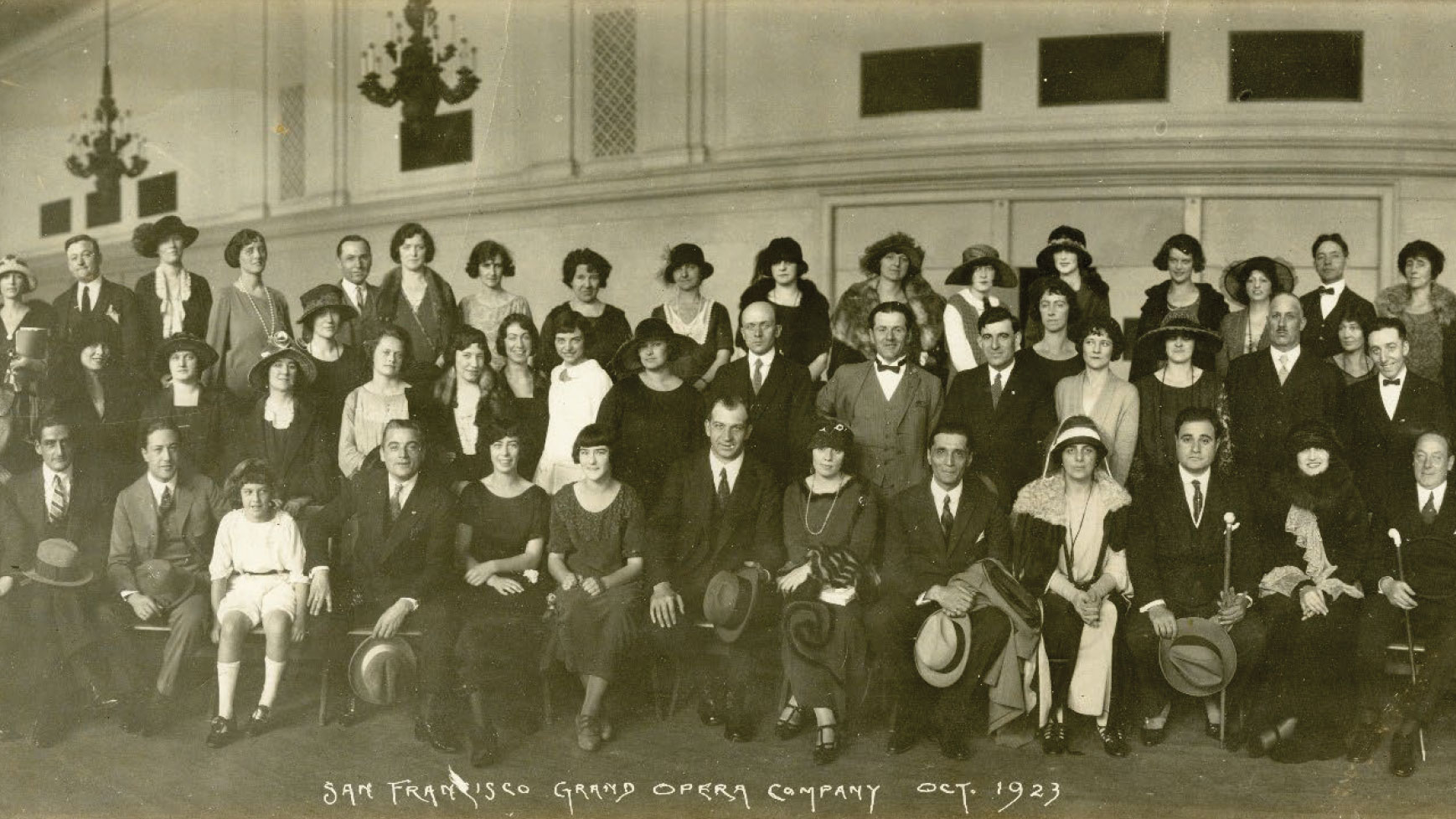 Large sepia-toned group photo of men, women and children on risers inside high-ceilinged space