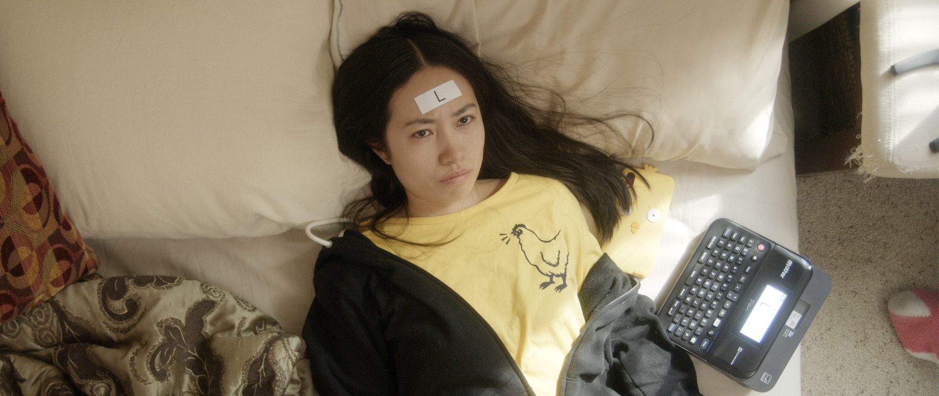 Person with "L" label on forehead lays on bed, label maker at side