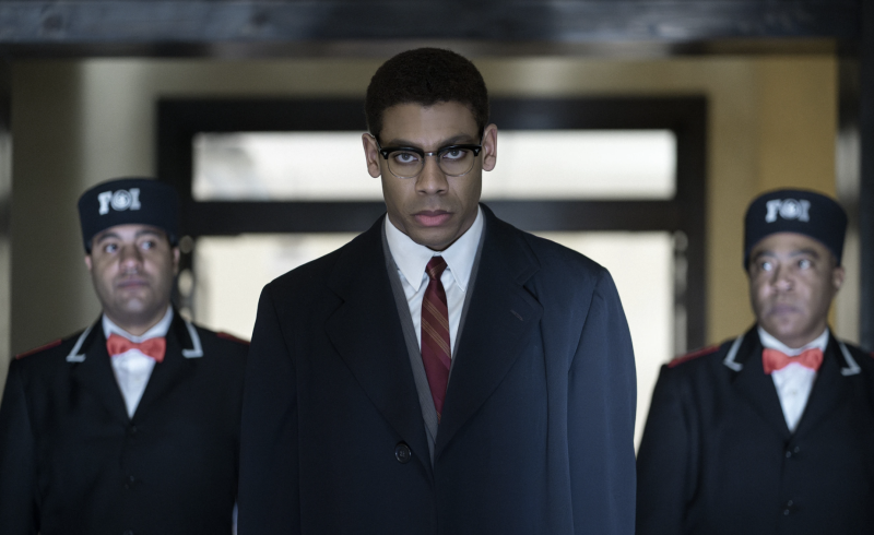 A Black man wearing spectacles and a 1950s-style suit and overcoat stands looking serious in a hallway. Two men wearing bowties stand behind him.