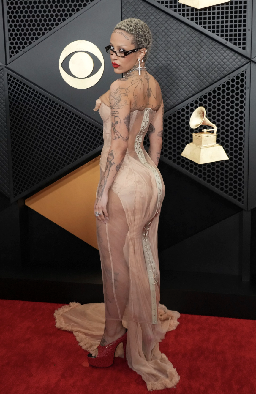 A tattooed woman stands in a sheer skin toned gown in the red carpet. She is looking over her shoulder.