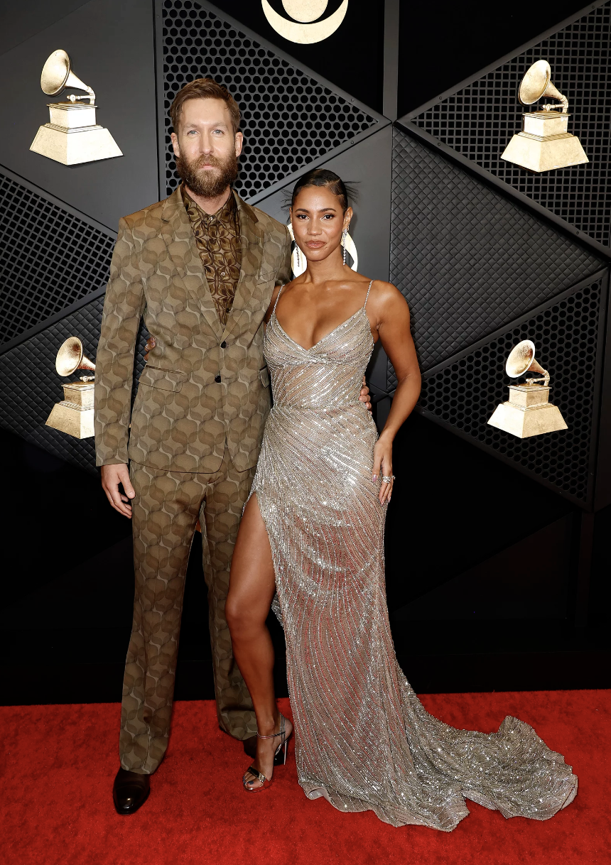 A man in a brown elaborately patterned suit stands with a woman in a high cut silver gown.
