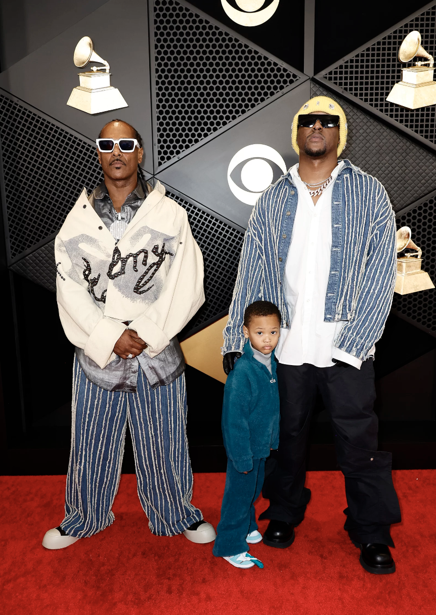 Two Black men wearing sunglasses and oversized shirts and pants stand side by side. One of them has a small boy at his side.
