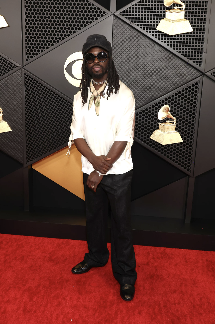 A Black man with shoulder length dreadlocks stands wearing a white shirt, black pants and sunglasses.