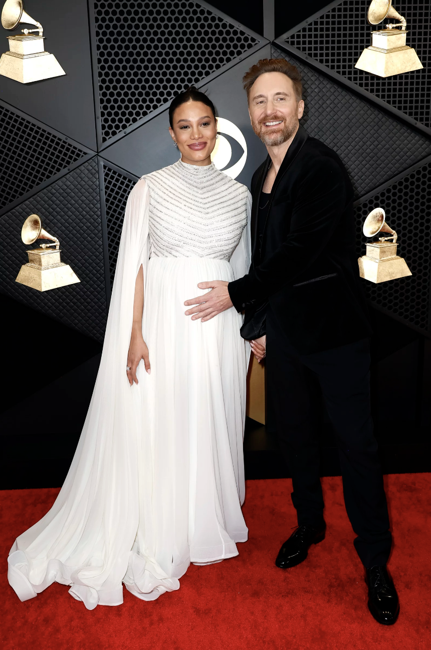 A man in a Black suit clutches the pregnant belly of a woman wearing a flowing white gown.