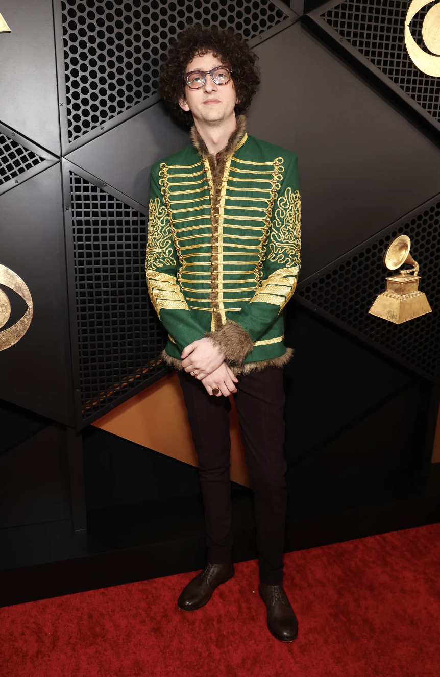 A white man with curly brown hair and glasses wearing black pants and green and gold band jacket.
