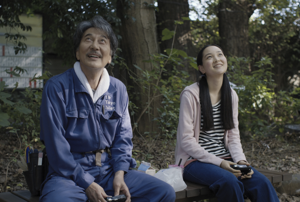 A Japanese man in blue overalls sits on a park bench next to a young teenage girl. They are both looking at the sky and smiling.