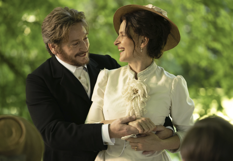 A man and a woman in period Victorian costume embrace and laugh together in a garden.