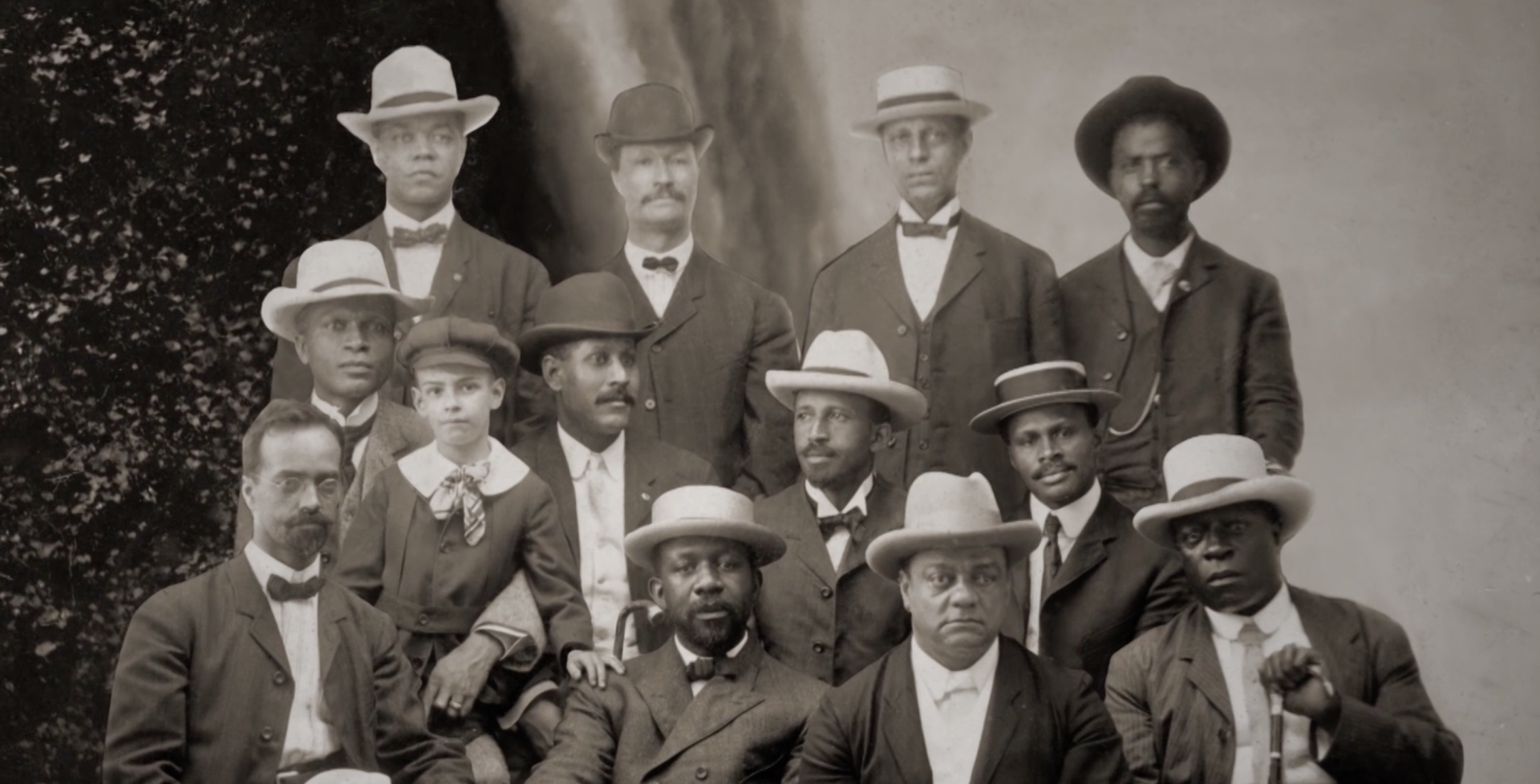 Twelve Black men and one Black boy, wearing turn-of-the-century clothing, sit in three rows for a photographic portrait.